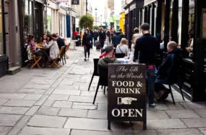 street view of people eating lunch. A majisign wooden a-board is in the foreground.
